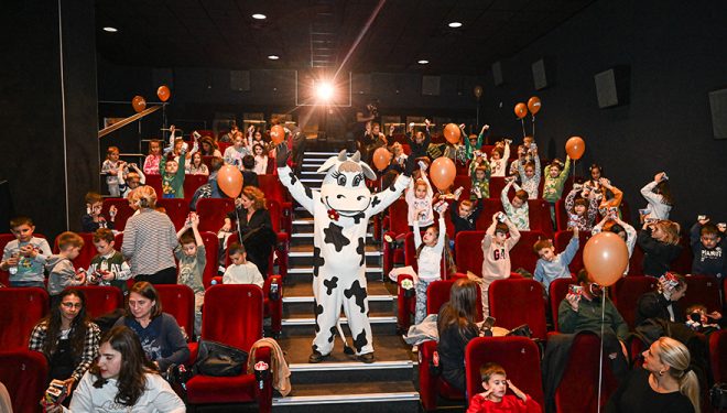 Maskota kravica raširenih ruku sa decom i roditeljima.